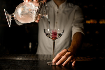 Professional bartender pouring a cocktail from the measuring cup with a strainer to a glass with pink ice cube