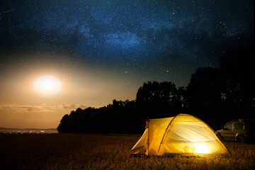 Fototapeta na wymiar Traveling and camping concept - camp tent at night under a sky full of stars. Orange illuminated tent and car. Beautiful nature - field, forest, plain. Moon and moonlight