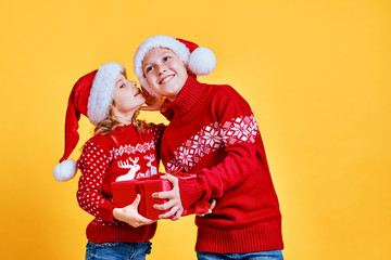 Fototapeta premium Little girl in red sweater with deer whispering secret to cheerful boy holding gift box on Christmas against yellow background