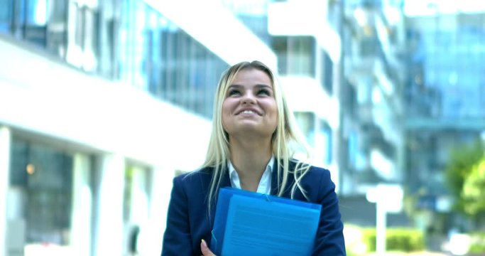 Holding a document folder and dressed in a white blouse and blue suit jacket, a blonde haired woman stands outside an office block, looking very excited