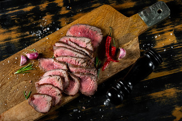 Beef meat steak sliced and served with herbs and spices on wooden cutting desk ob wooden black background.