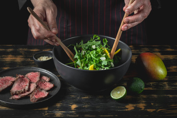 Cooking fresh diet keto salad with beef meat, arugula, fruits and nuts by chef hands on black table background.