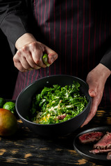 Cooking fresh diet keto salad with beef meat, arugula, fruits and nuts by chef hands on black table background.