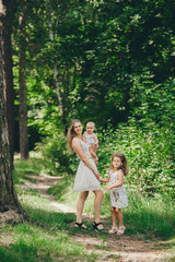 Naklejka na ściany i meble Mother and two little daughter in summer dresses go through the woods in the summer. Mother and children outdoors