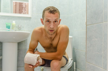 A dark-haired man in shorts with toilet paper in his hands sits on the toilet and looks at the...