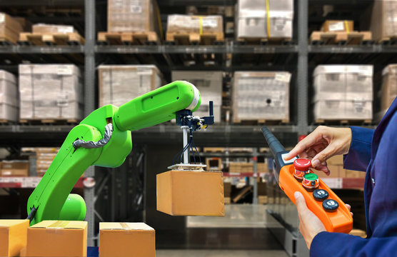 Industrial Robot Holding A Box And Worker Operating A Robot Machine With A Control Panel On Stock Shelves Background