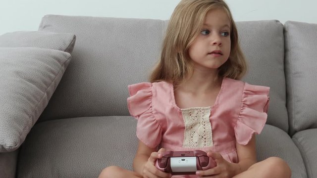 Anxious preteen girl in earphones sitting on sofa, holding joystick and paying video game 4k