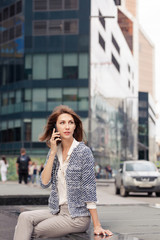Fototapeta premium beautiful young business brunette woman dressed in elegant clothes sits on a bench on a street in the city center and talking on a cell phone. Glass building in the background