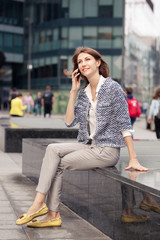 Fototapeta premium attractive smiling young business brunette girl dressed in elegant clothes sits on a bench on a street in the city center and talking on mobile phone. Glass building in the background