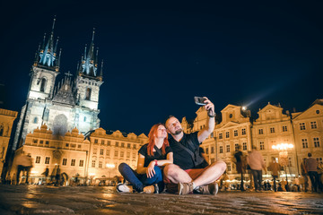 Loving couple sit in the main square, taking pictures on the phone, selfie smartphone, happy together, on the street of the old city of Prague, night time. historical center. travel and tour in Europe