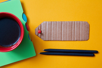 Obraz premium A red Cup of coffee and a notebook on a yellow background and a cardboard sign for the inscription. Mock up.