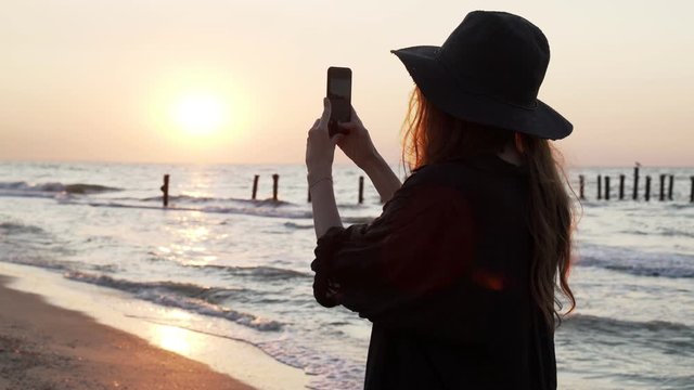 Girl in a black hat filming beautiful sunset on a seaside. Summer warm golden hour light, evening landscape and sea. Yellow sand and calm waves. Woman making selfie? photos and videos on telephone 