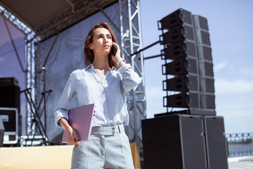Installation of stage equipment and preparing for a live concert open air. Event manager portrait. Summer music city festival. Young serious woman stand and work with her laptop near the stage... - 287022619