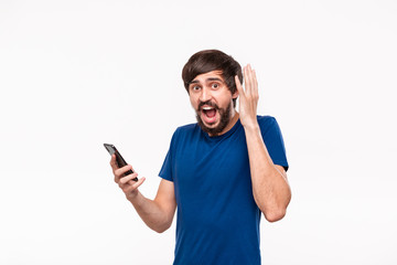 Pretty brunet man in a blue shirt with beard and mostaches shocked holding smartphone looking at the camera standing isolated over white background.