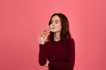 Excited pretty brunette girl drinking sparkling champagne from a wineglass standing isolated on a dark pink background.