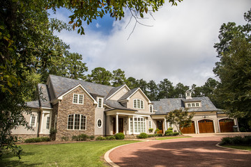 Front view of large estate home in the south with a gravel driveway and lots of windows. house made of brick, stone and clapboard in a cape cod style. and a triple garage with curb appeal