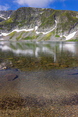 Flock of small fish, ripple in the clean, transparent water of famous Kidney lake, one of seven Rila lakes, epic, sunlit rocky summits and a blue sky with white clouds