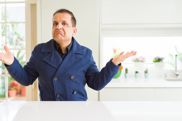 Middle age man sitting at home clueless and confused expression with arms and hands raised. Doubt...