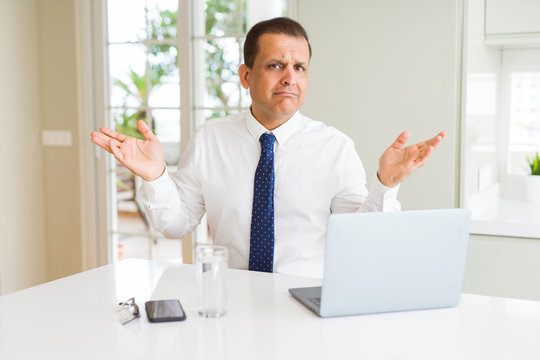 Middle age business man working with computer laptop clueless and confused expression with arms and hands raised. Doubt concept.
