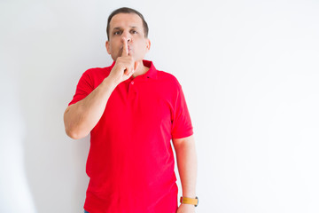 Middle age man wearing red t-shirt over white wall asking to be quiet with finger on lips. Silence and secret concept.