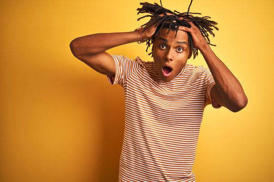 Afro man with dreadlocks wearing striped t-shirt standing over isolated yellow background Crazy and scared with hands on head, afraid and surprised of shock with open mouth