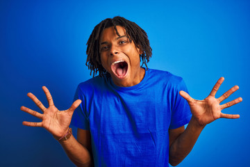 Afro american man with dreadlocks wearing t-shirt standing over isolated blue background celebrating crazy and amazed for success with arms raised and open eyes screaming excited. Winner concept