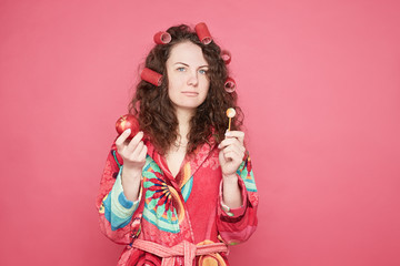Obraz na płótnie Canvas Studio shot of thoughtful pretty indecisive curly haired housewife with curlers on head feeling confused and hesitant, facing difficult choice: whether to eat healthy organic apple or unhealthy candy.