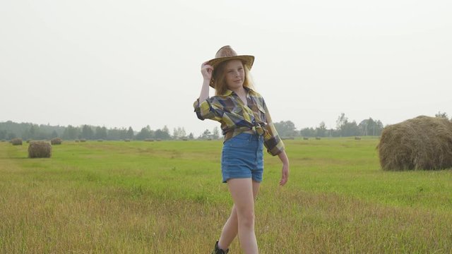 Country Style Little Girl Walking on Green Field. Summer Countryside Vacation. Caucasian Young Lady in Cowboy Straw Hat. Blond Farmer Teenage Daughter with Freckles on Haystack Landscape