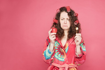 .Studio shot of pretty housewife in colorful bathrobe feeling confused and hesitant, facing difficult choice: whether to eat healthy organic apple or unhealthy candy, isolated on pink with copy space.
