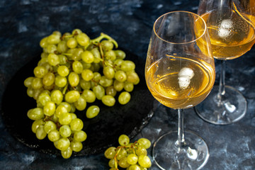 White grapes lie on a black plate. Nearby are two glasses filled with rose wine. Ice cubes for decoration. All on a dark background.