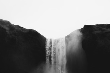Falling water of Skogarfoss waterfall between green hills in Iceland. Gray cloudy sky and white splashes. Gothic black and white tones.