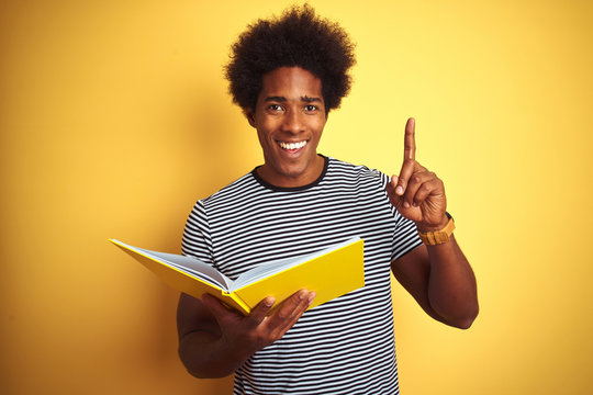 Afro american student man reading book standing over isolated yellow background surprised with an idea or question pointing finger with happy face, number one