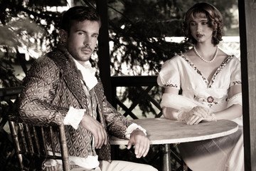 Handsome young couple in vintage clothing sit in gazebo with handsome gentleman looking at camera as companion looks on.