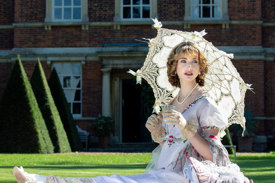 Beautiful lady in regency clothing sitting on lawn in front of stately home holding parasol and drinking sparkling wine
