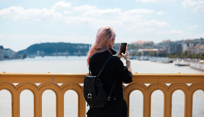 Young girl with backpack and pink hair, take a photo with smartphone from the bridge.