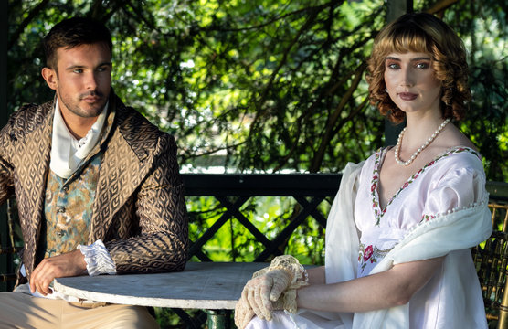 Handsome young couple in vintage clothing sit in gazebo. The female is looking at camera as her companion looks at her.