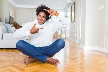 African American man sitting on the floor at home smiling making frame with hands and fingers with happy face. Creativity and photography concept.