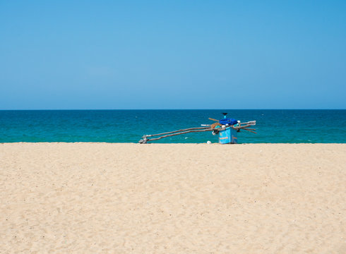 Traditional wooden fishing boat on empty beach against clear blue sky and sea, Kallady Beach, Batticaloa, Sri Lanka.