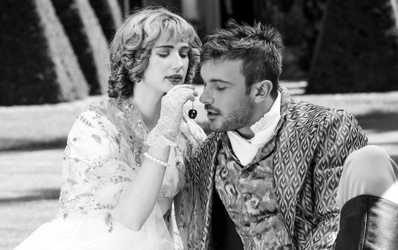 Young lovers dressed in vintage clothing. Man looks on as his partner feeds him a cherry