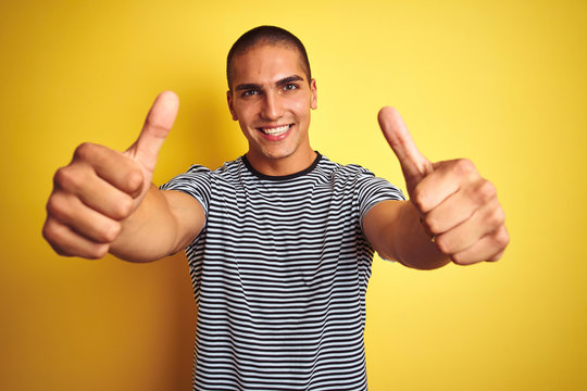 Young handsome man wearing striped t-shirt over yellow isolated background approving doing positive gesture with hand, thumbs up smiling and happy for success. Winner gesture.
