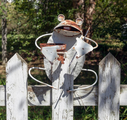 Rusty yet cute metal frog ornament holding empty flower pot atop white picket fence.