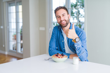 Handsome man eating cereals for breakfast at home doing happy thumbs up gesture with hand. Approving expression looking at the camera with showing success.