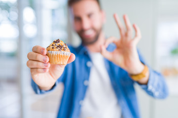 Handsome man eating chocolate chips muffin doing ok sign with fingers, excellent symbol