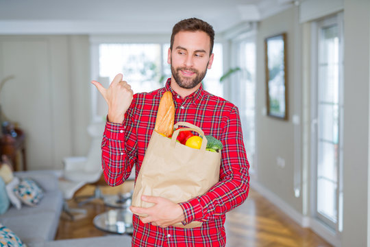 Handsome man holding groceries bag pointing and showing with thumb up to the side with happy face smiling
