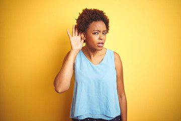 Beautiful african american woman wearing elegant shirt over isolated yellow background smiling with hand over ear listening an hearing to rumor or gossip. Deafness concept.