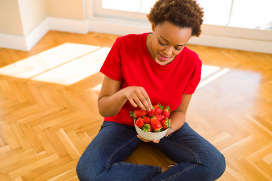 Beautiful young african woman with afro hair eating fresh strawberries sitting on the floor