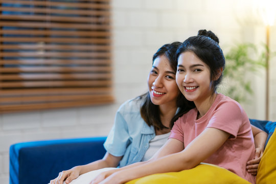LGBTQ asian lasbian couple smiling ,looking at camera and sitting on sofa at living room at house. Asian woman and friend feeling happy in evening time. LGBT and friendship concept.