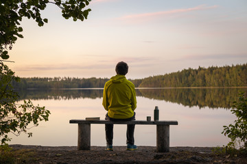 A man in a yellow jacket sits on a bench by the lake on a calm autumn evening. Near a thermos and a...