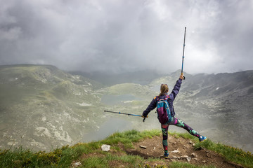A girl with colorful tights, backpack and walking sticks standing on the edge of Lake peak on Rila mountain, famous viewpoint of seven Rila lakes and looking at iconic lakes through the mist