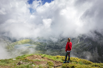 A girl with red jacket and colorful tights standing on the edge of Lake peak on Rila mountain, famous viewpoint of seven Rila lakes and looking at iconic lakes through the dramatic mist